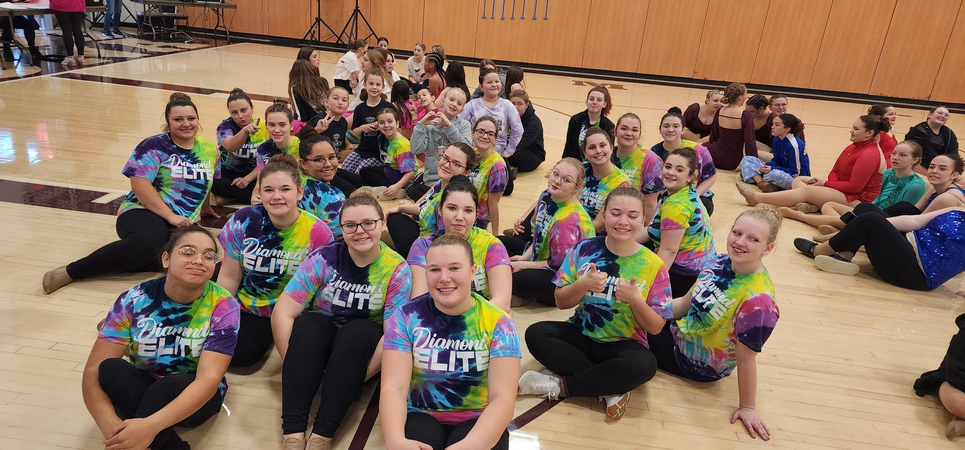 Group of people in tie-dye shirts sitting on a gym floor, smiling and posing.