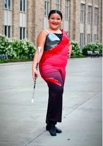 Young person in baton twirling uniform poses outside, smiling, holding baton. Bright colors on costume.