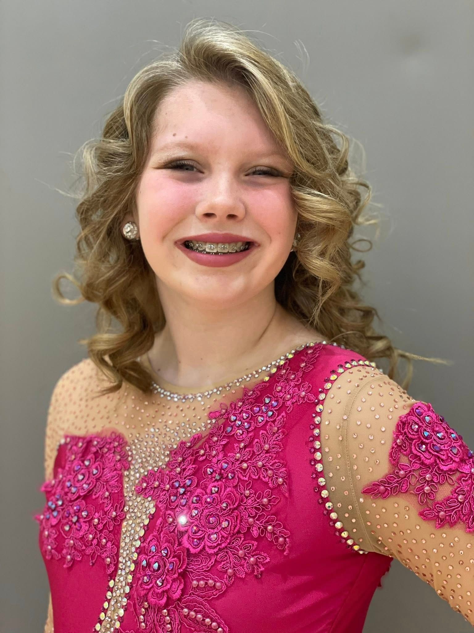 Woman wearing a pink embellished dress smiles, has curly blonde hair, and braces.