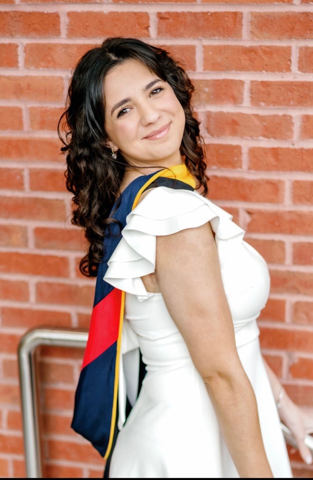 Woman in a white dress, smiling, with graduation sash, leaning against a brick wall.