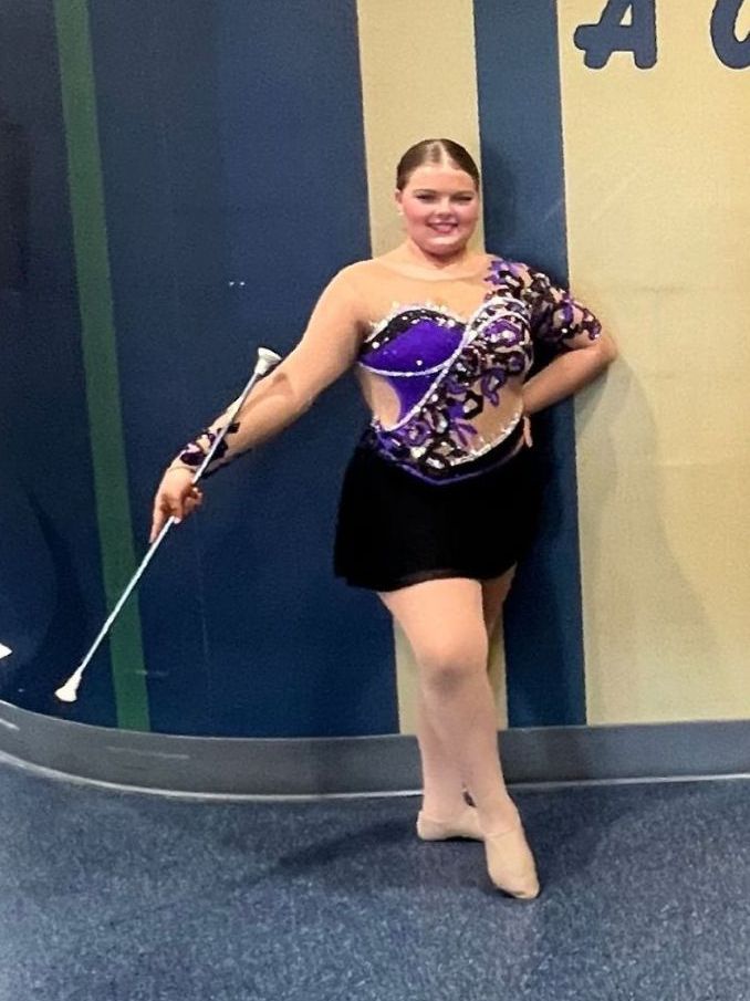Woman in purple and black costume twirling batons, posing against a blue and gold wall.