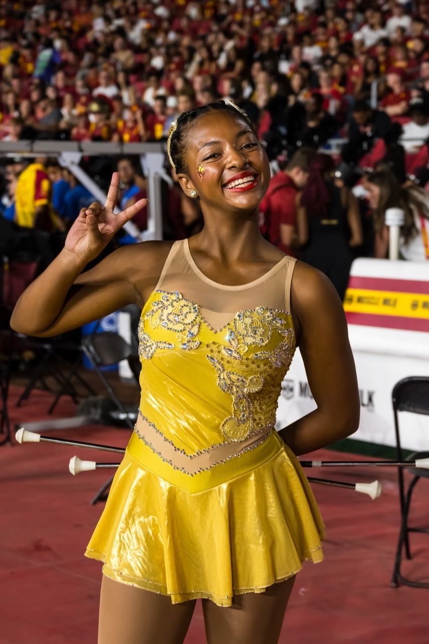Woman in yellow costume with baton, stadium crowd background, giving peace sign.