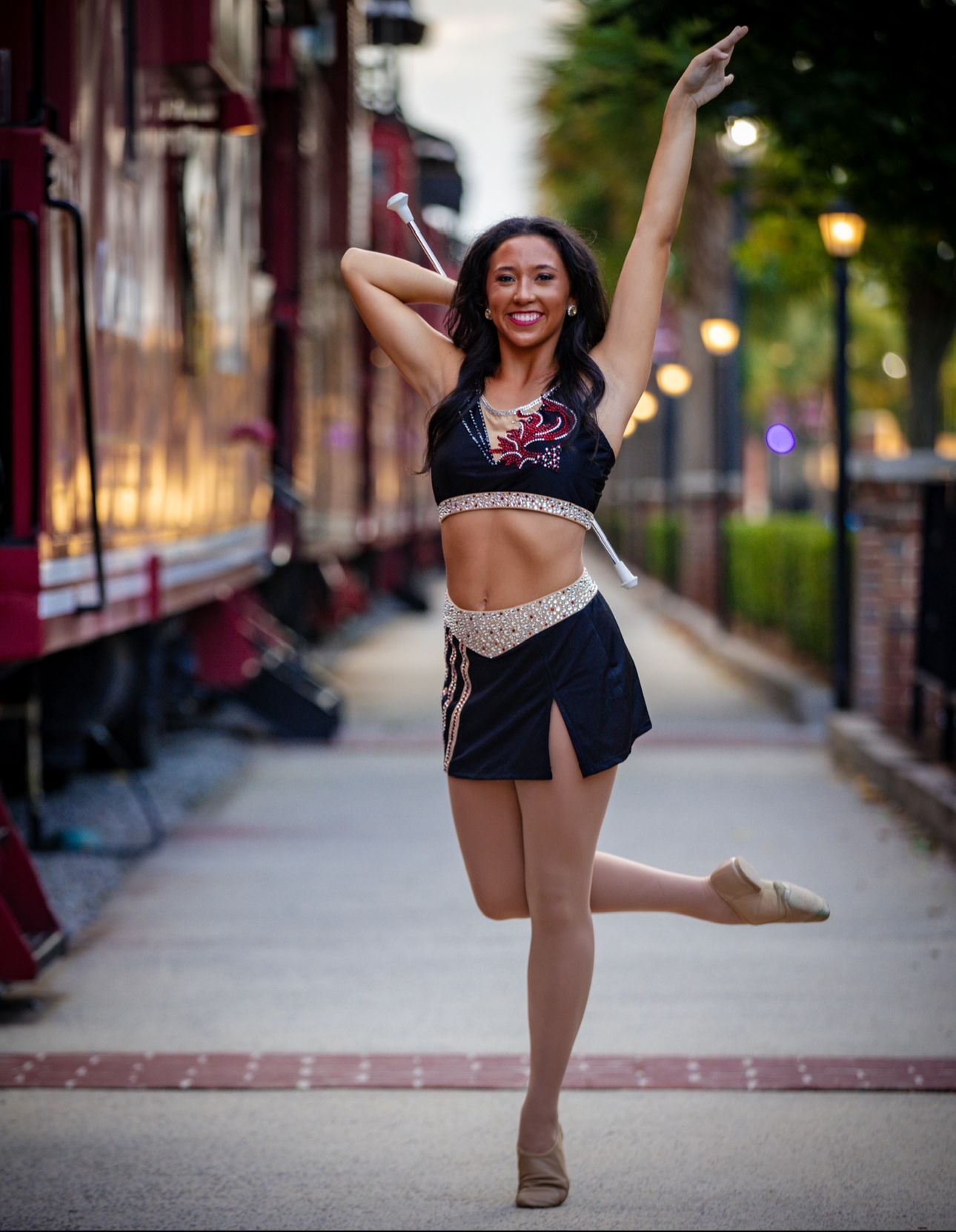 Woman twirling baton, wearing black and gold outfit, posing next to train.
