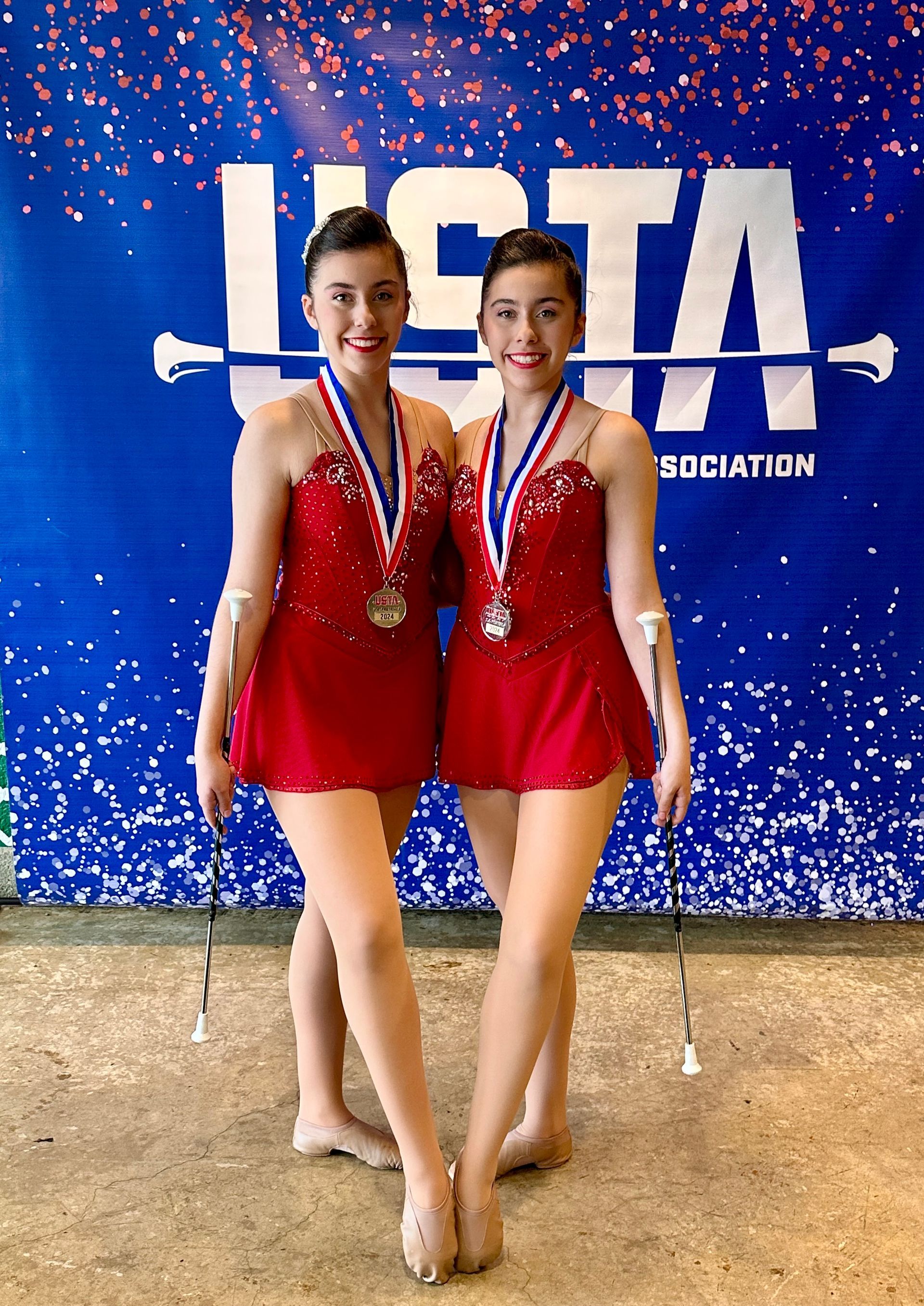 Two women in red dresses with batons and medals posing in front of a blue banner.