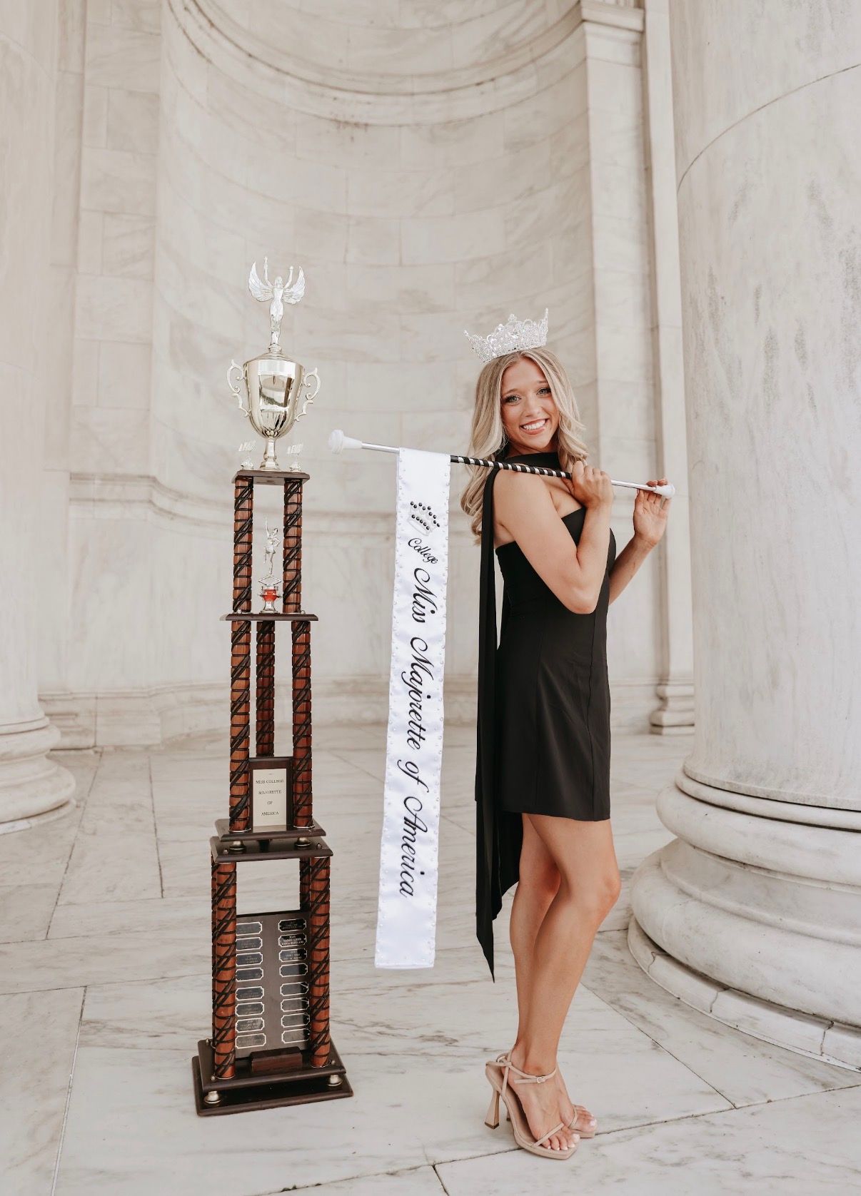 Woman in a black dress posing beside a trophy in a marble-columned hall
