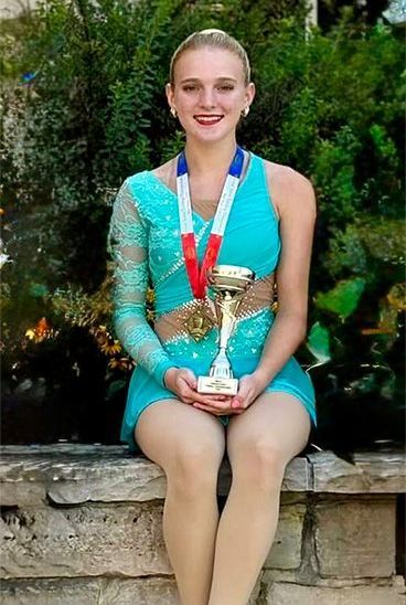 Woman in a teal skating costume, holding trophy and medals, smiling. Sitting outdoors.