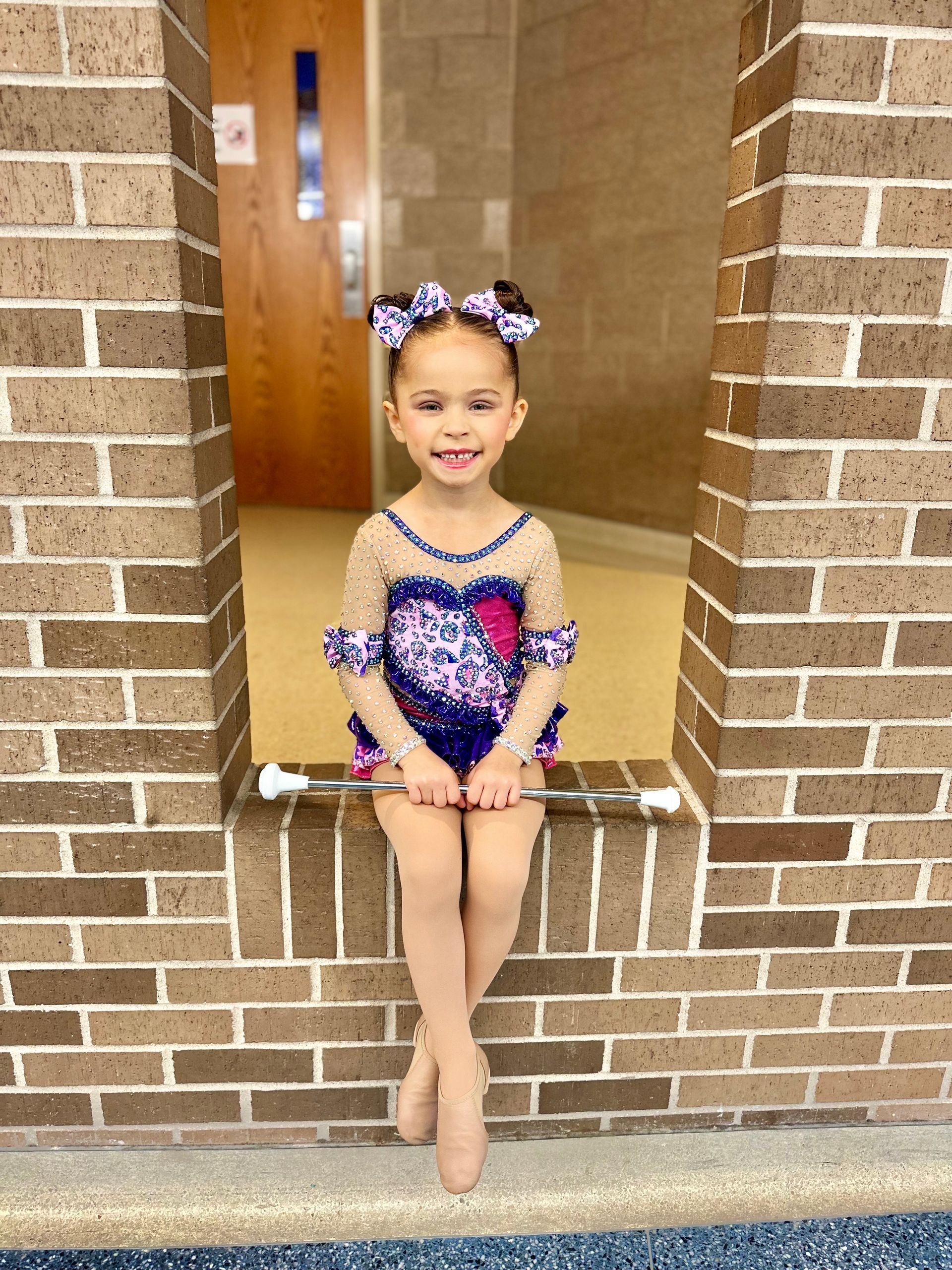 Young girl in baton twirling costume poses in brick alcove, holding baton, smiling.