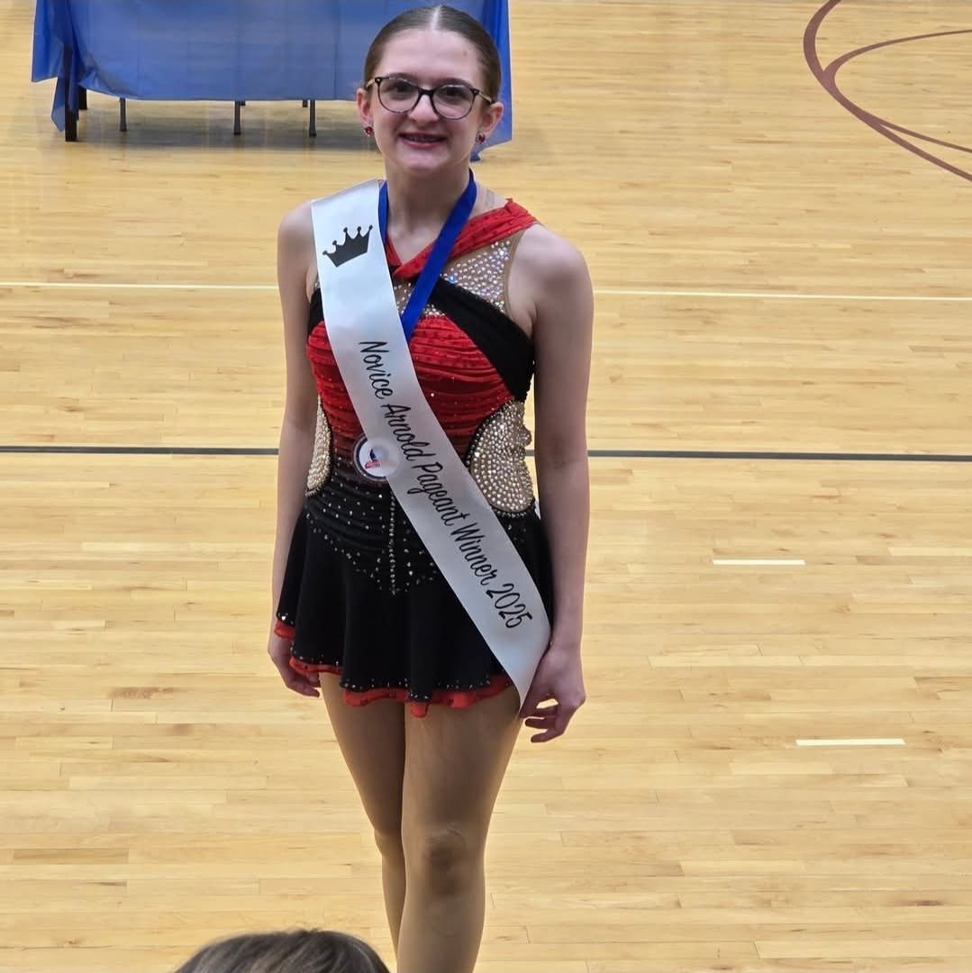Young person wearing a sash and crown, in a competition leotard, stands on a gym floor.