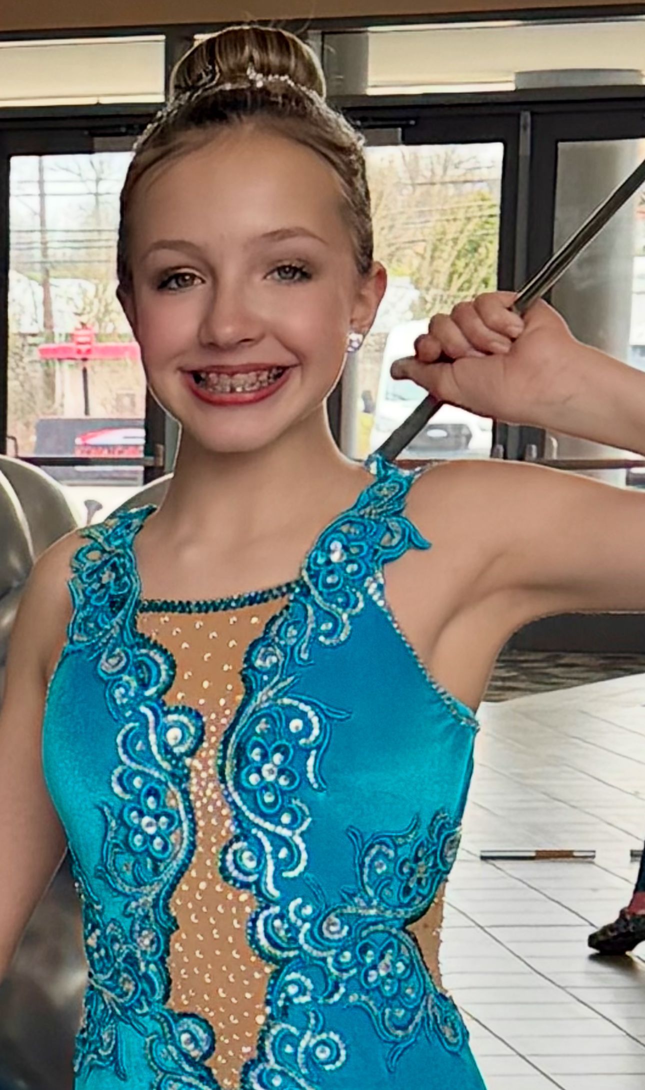 Young person in a teal costume, holding a baton, smiling, with braces and a bun.