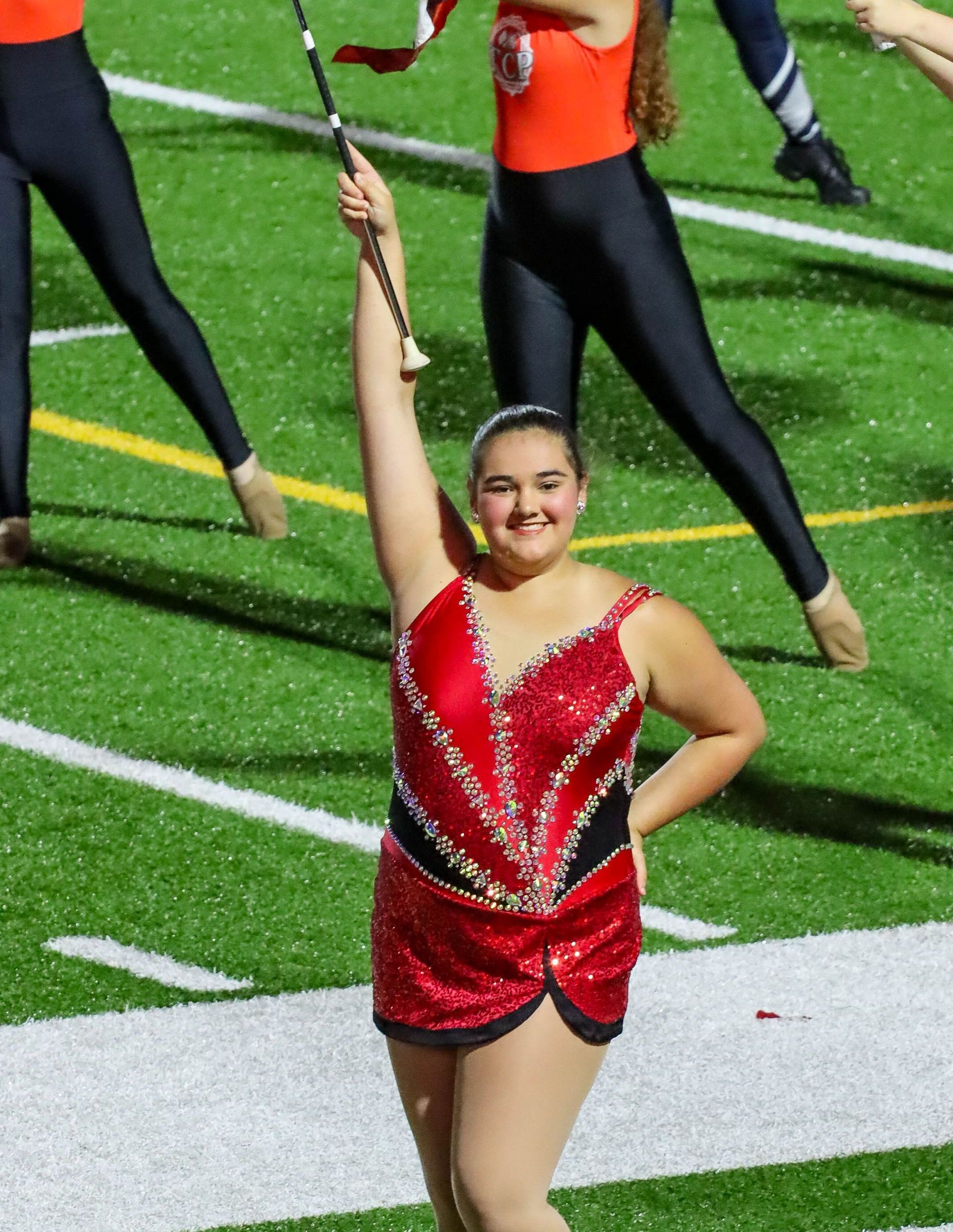 A baton twirler in a red costume with a raised baton on a green field.