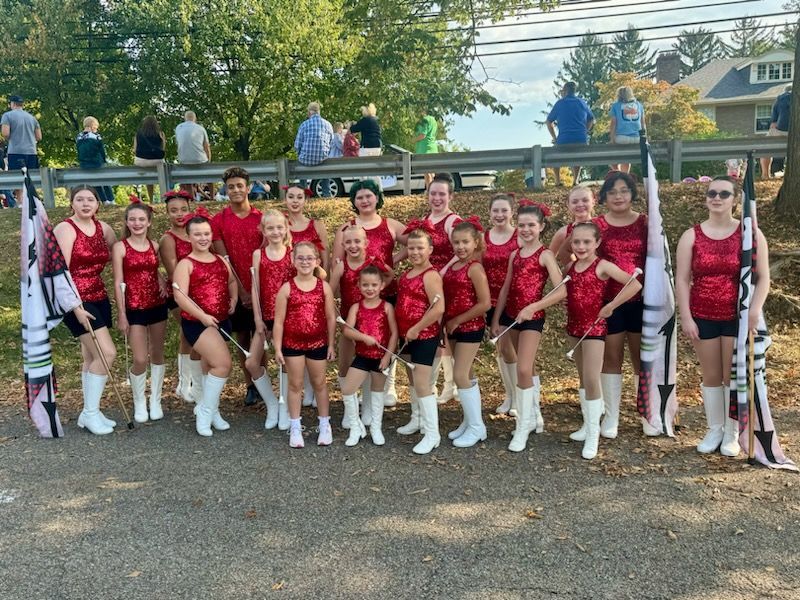 Group of twirlers in red sequin tops, black shorts, and white boots, posing with flags outdoors.