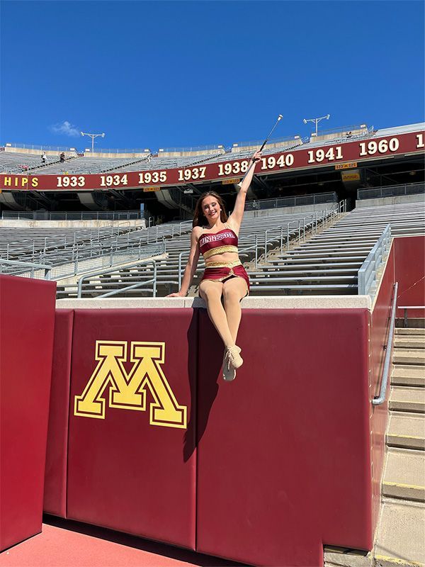 A person in maroon and gold attire twirling a baton, posing on a wall with a