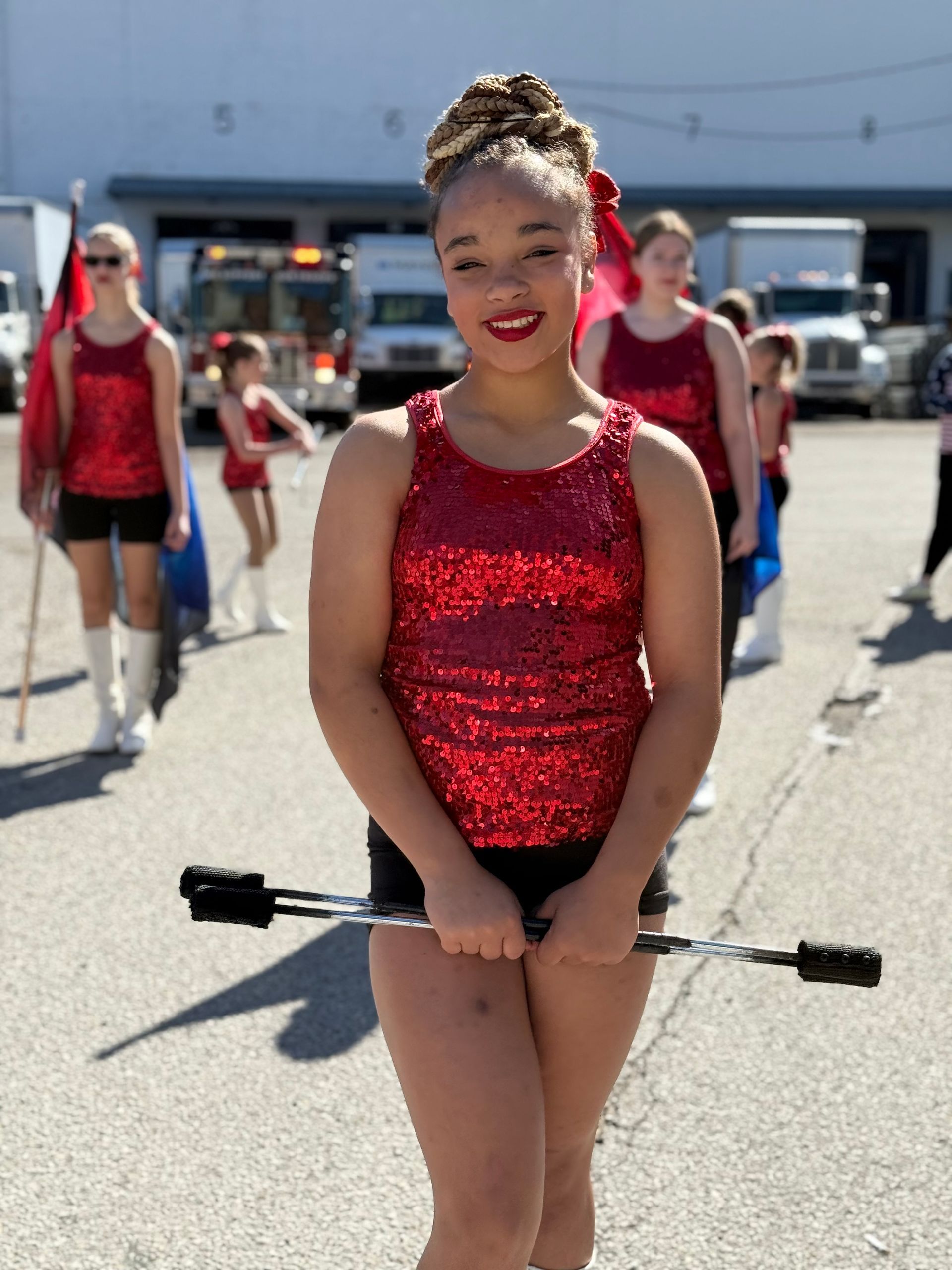 Girl in red sequin top holding a baton, part of a marching group outdoors.