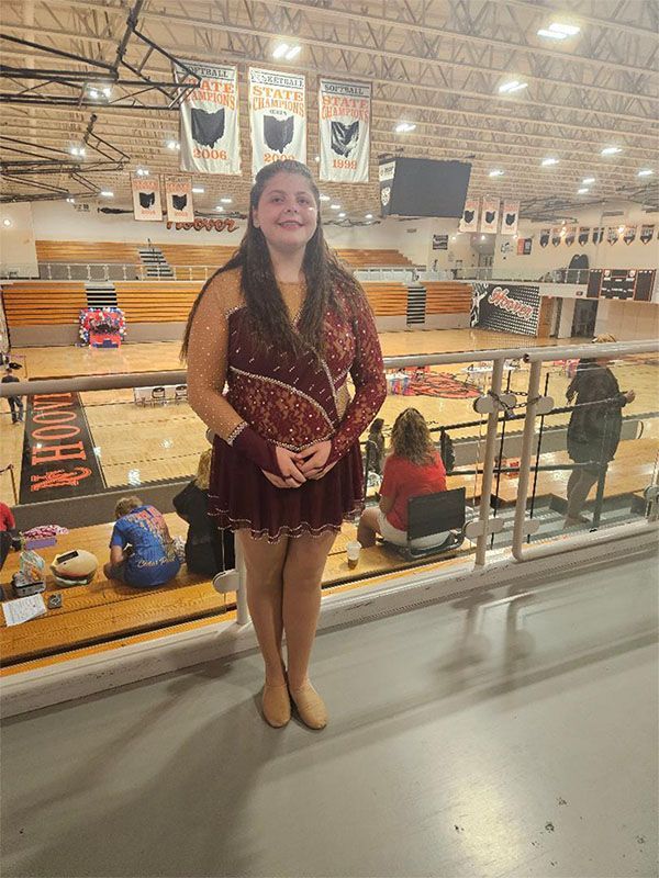 Person in a maroon dance costume stands in an indoor gym with a basketball court.