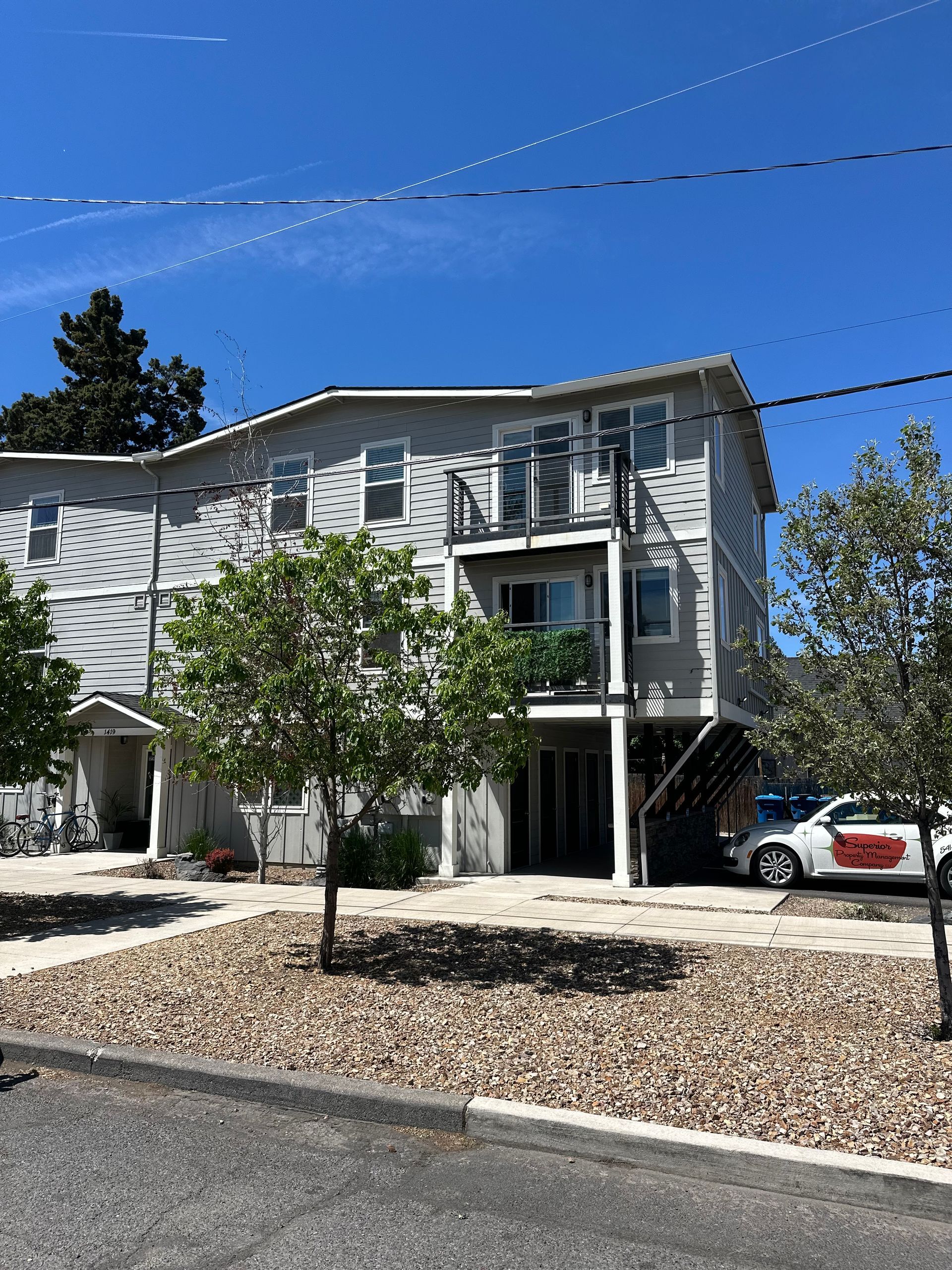 White two-story house with balconies, trees, and a parked car under a clear blue sky