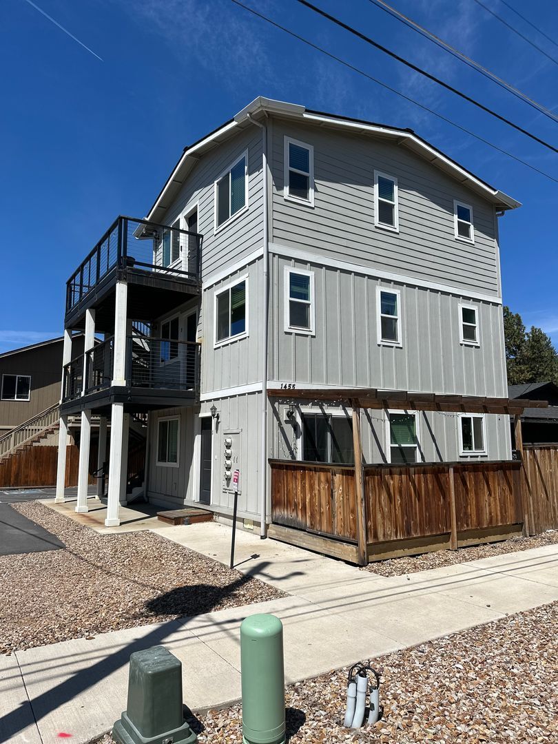 Three-story gray house with white trim and a wooden deck under construction on a sunny street corner