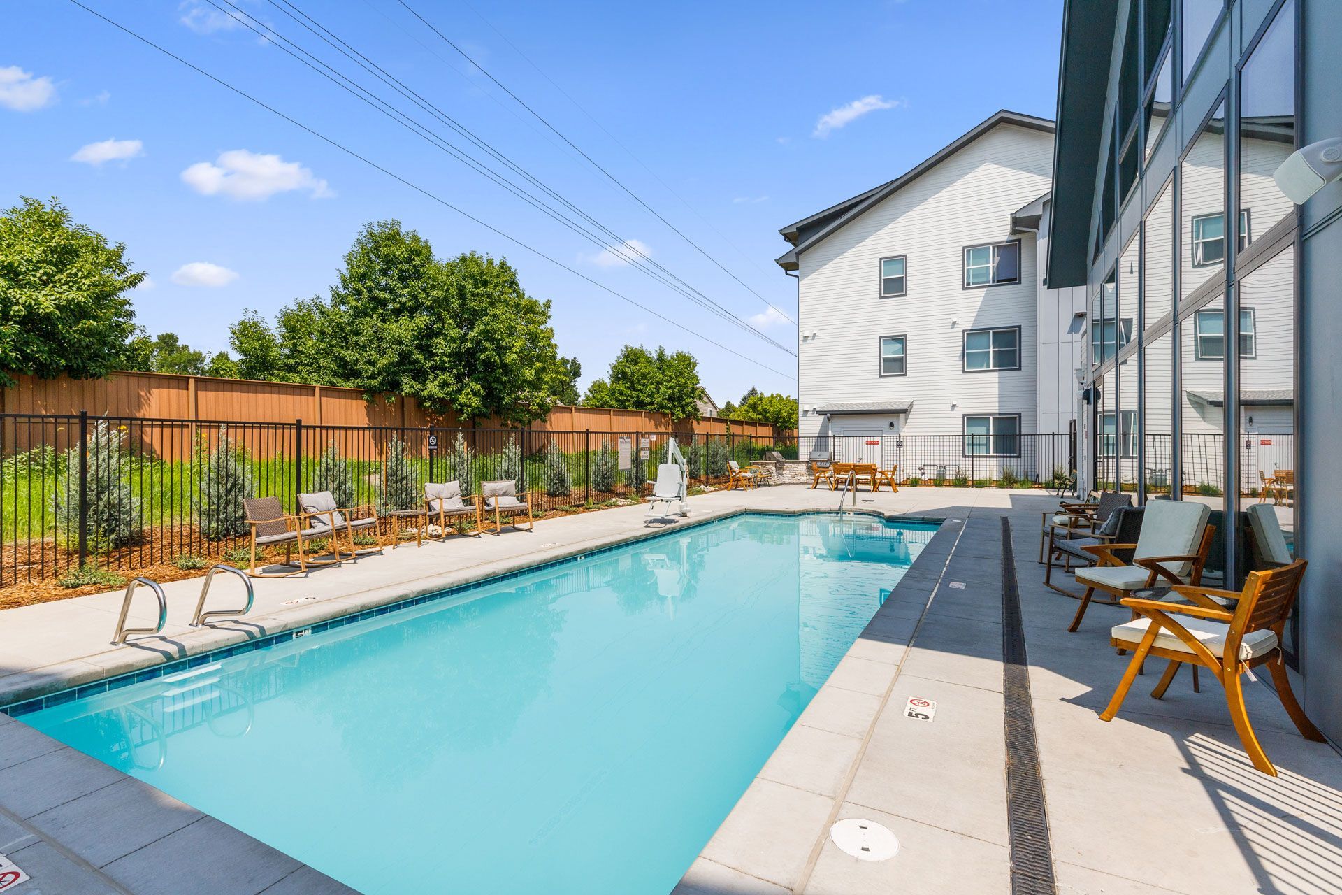 An outdoor swimming pool at a multi-story building with a wooden fence and trees in the background on a sunny day.