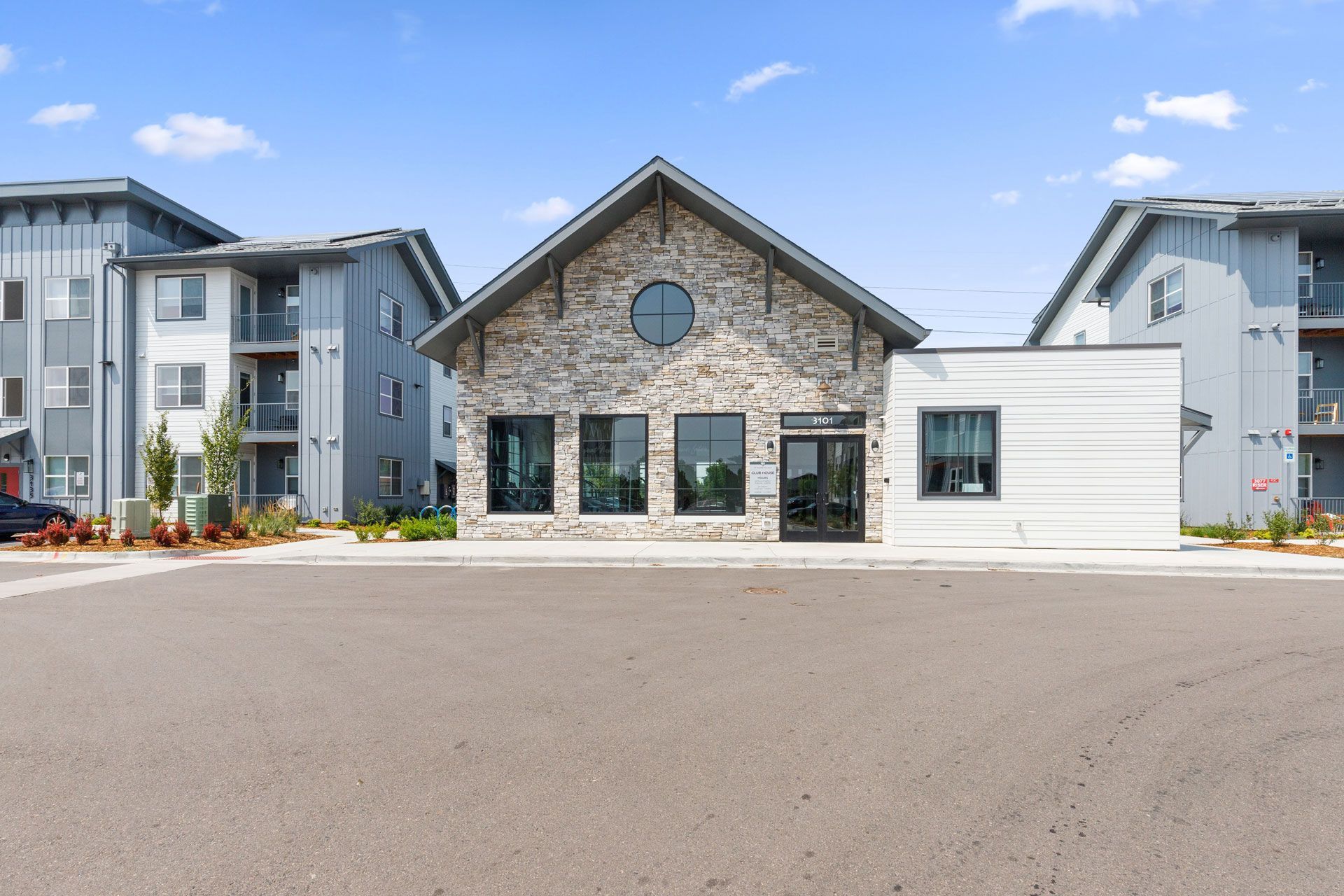 A stone-front clubhouse building sits between two modern multi-story apartment buildings under a clear blue sky.