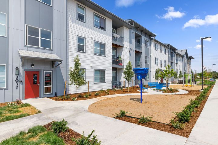 A modern multi-story apartment building with a red entrance door, white siding, and a small play area in the front yard.