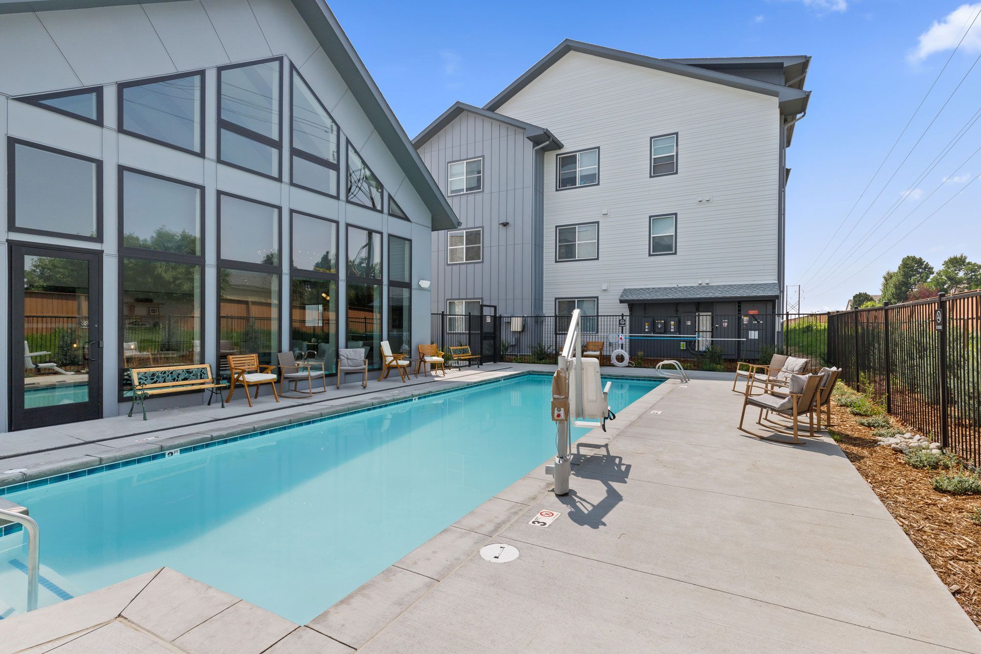 A bright outdoor swimming pool sits next to a modern building with large floor-to-ceiling windows and patio furniture.