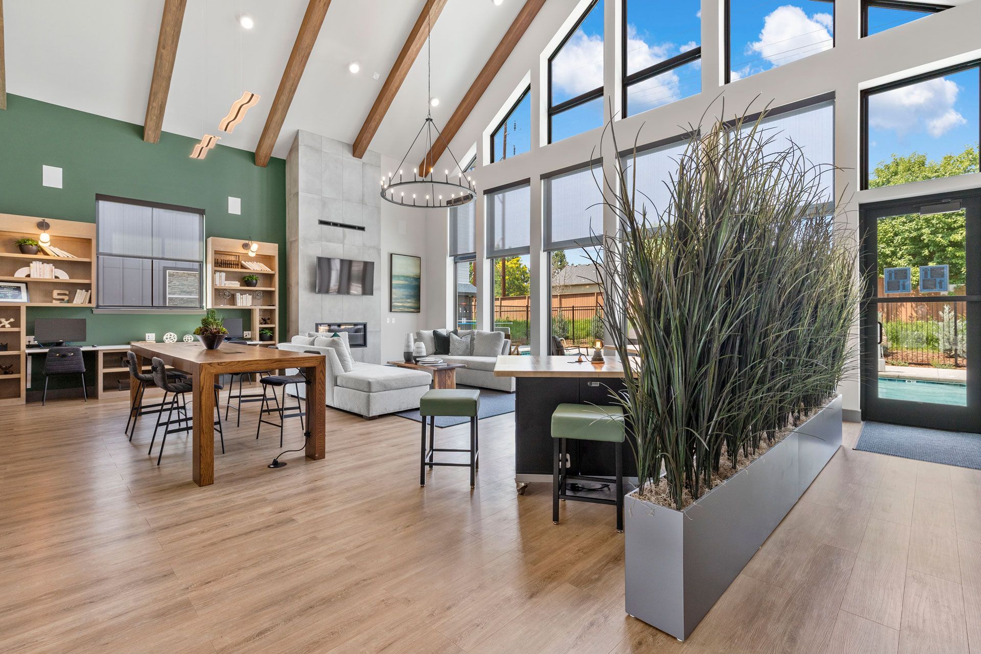A bright modern community lounge with wood floors, vaulted ceilings, a large dining table, seating, and indoor greenery.