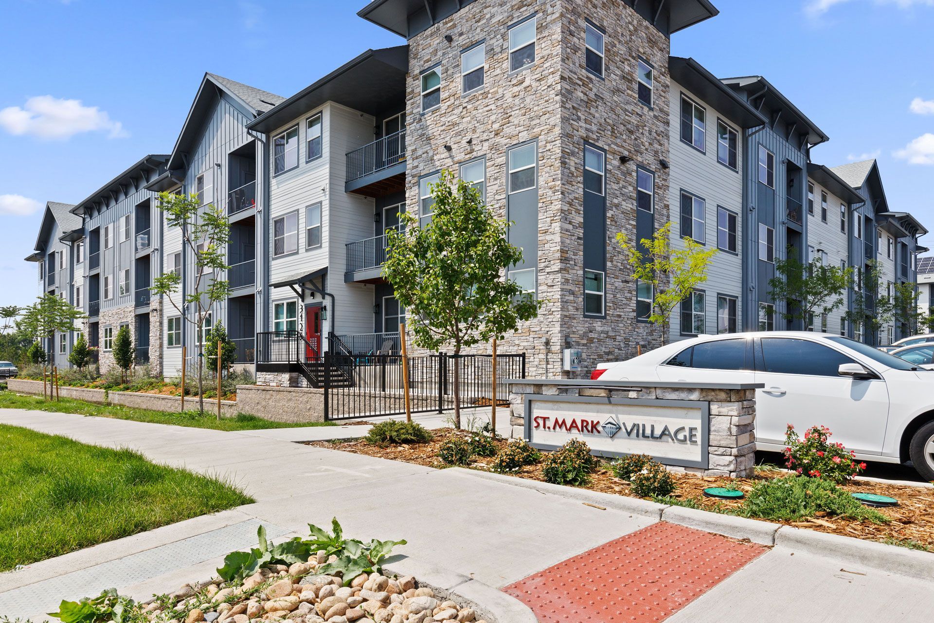 Multi-story apartment building with stone and neutral siding, next to a sidewalk and a sign reading 