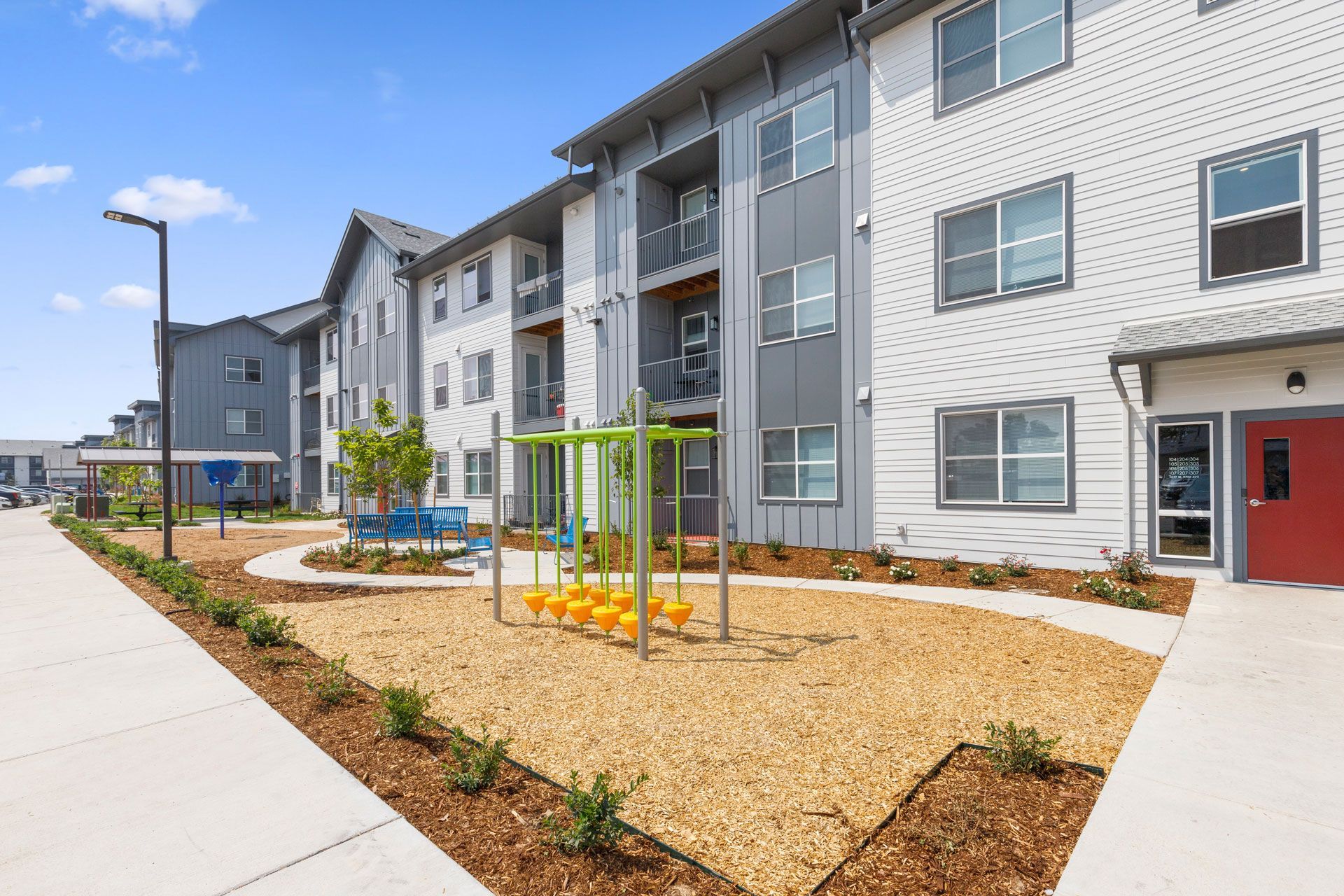 A sunny apartment complex exterior with a small playground area featuring yellow climbing equipment on wood chips.