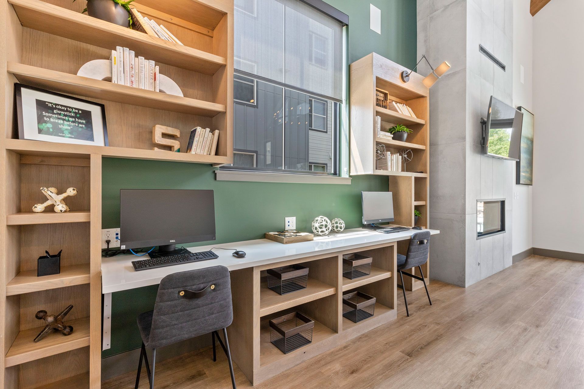 A bright home office with two workstations, light wood shelving, a green accent wall, and a fireplace to the right.