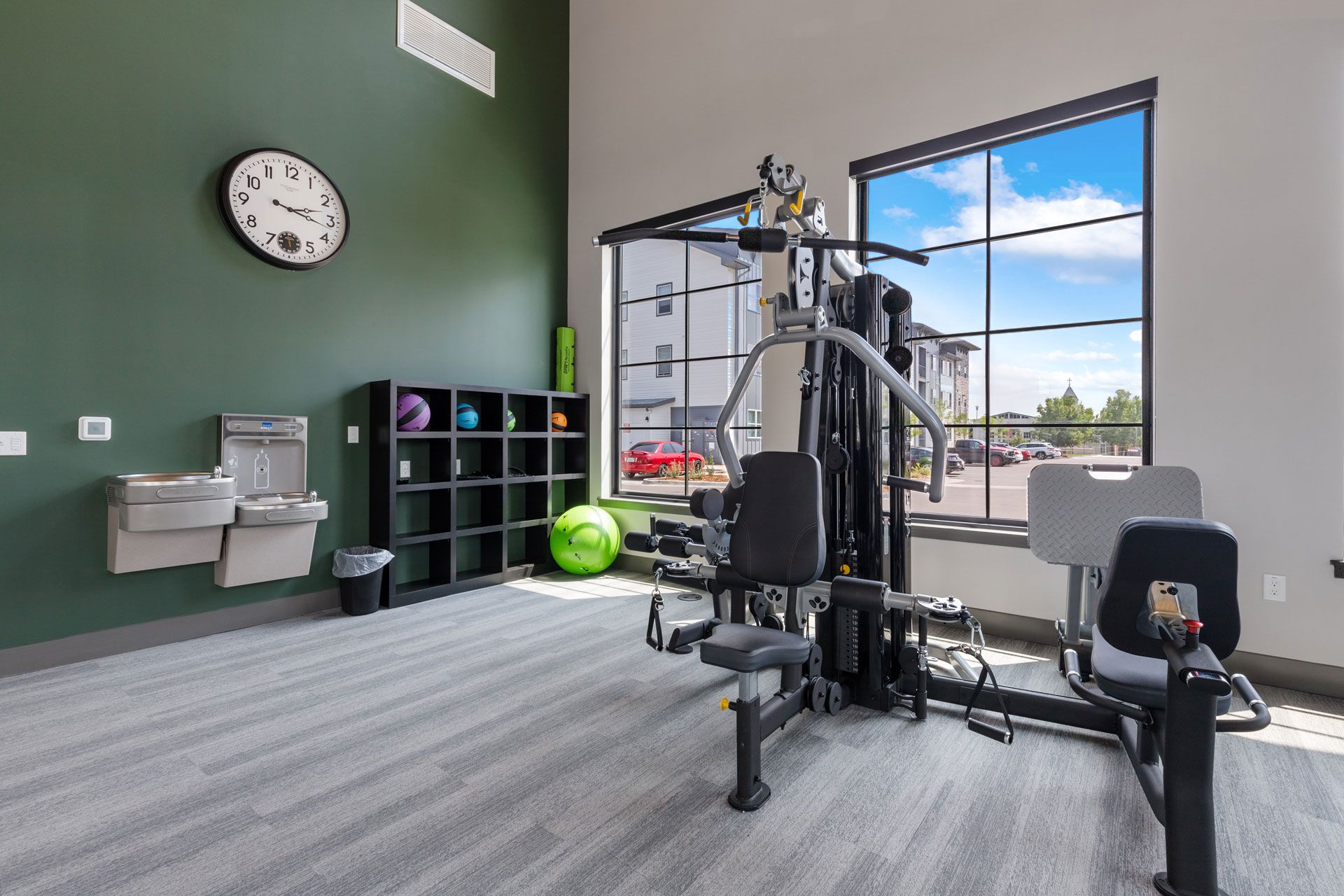 A gym with a large weight machine, a water fountain, a black storage unit with exercise balls, and a clock on a green wall.