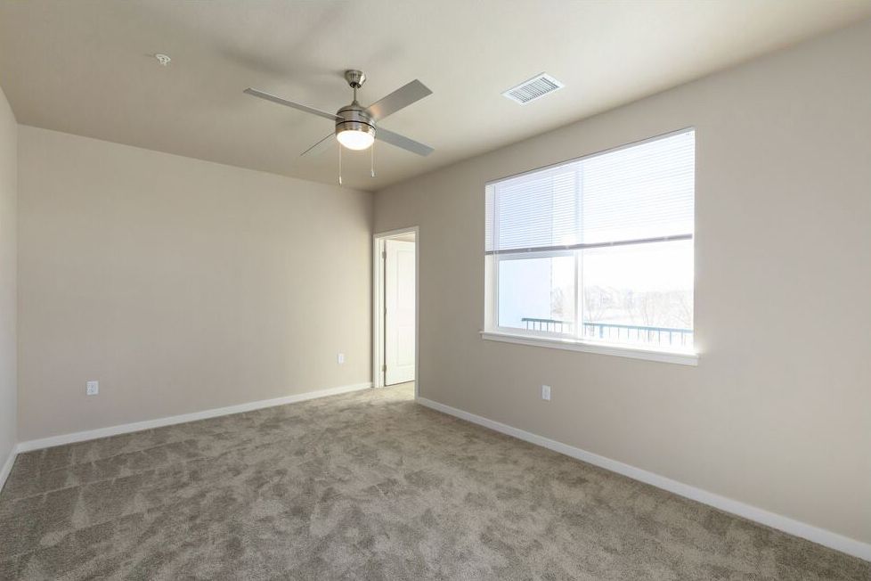 Empty bedroom with beige walls, gray carpet, a ceiling fan, and a window with white blinds.