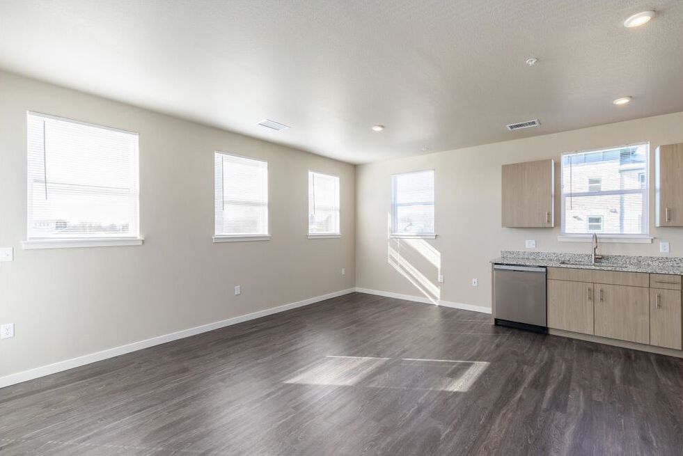 Modern, minimalist living space with light walls, dark wood-look flooring, multiple windows, and a small kitchen area.