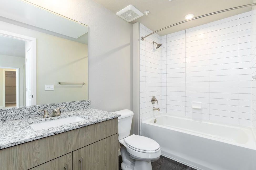 A modern bathroom featuring a granite-topped vanity, white tiled shower and tub combo, and a standard toilet.