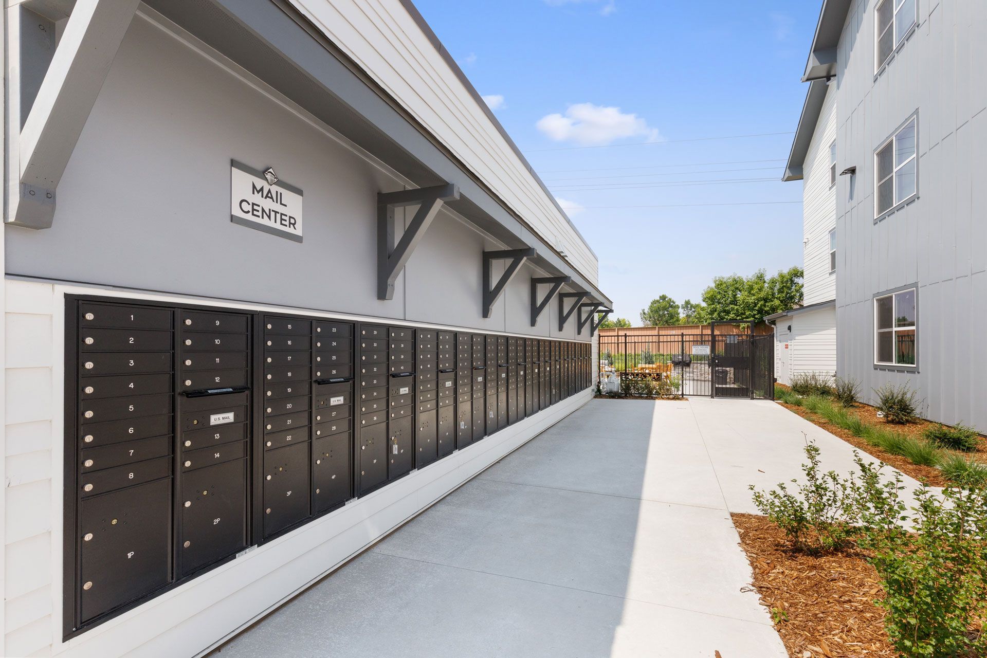 A row of black mailboxes mounted on a gray wall outdoors under a clear blue sky, with a concrete walkway alongside.