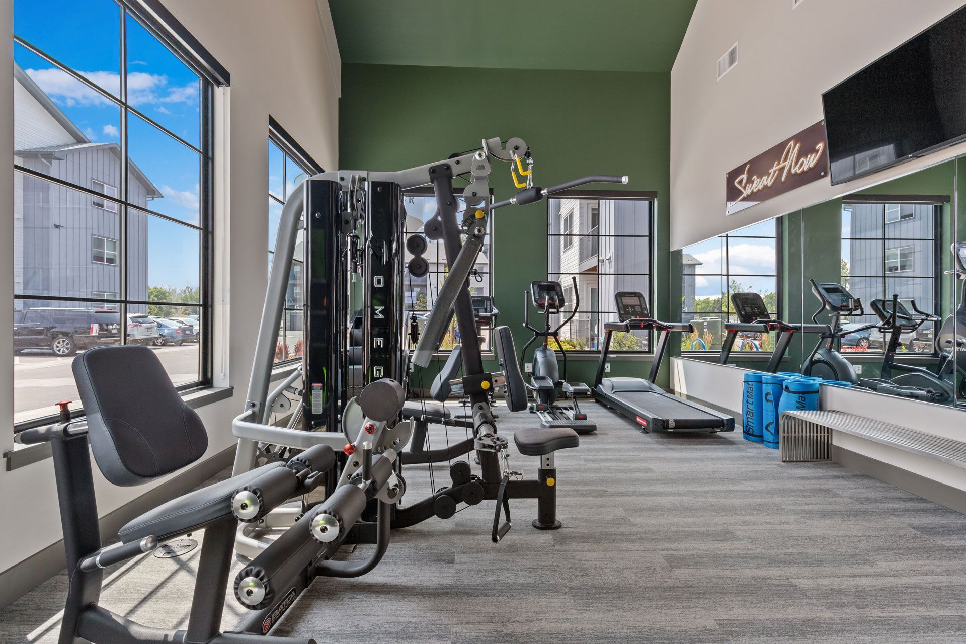 A bright gym with a multi-station weight machine, treadmill, and stationary bike, featuring large windows and green walls.