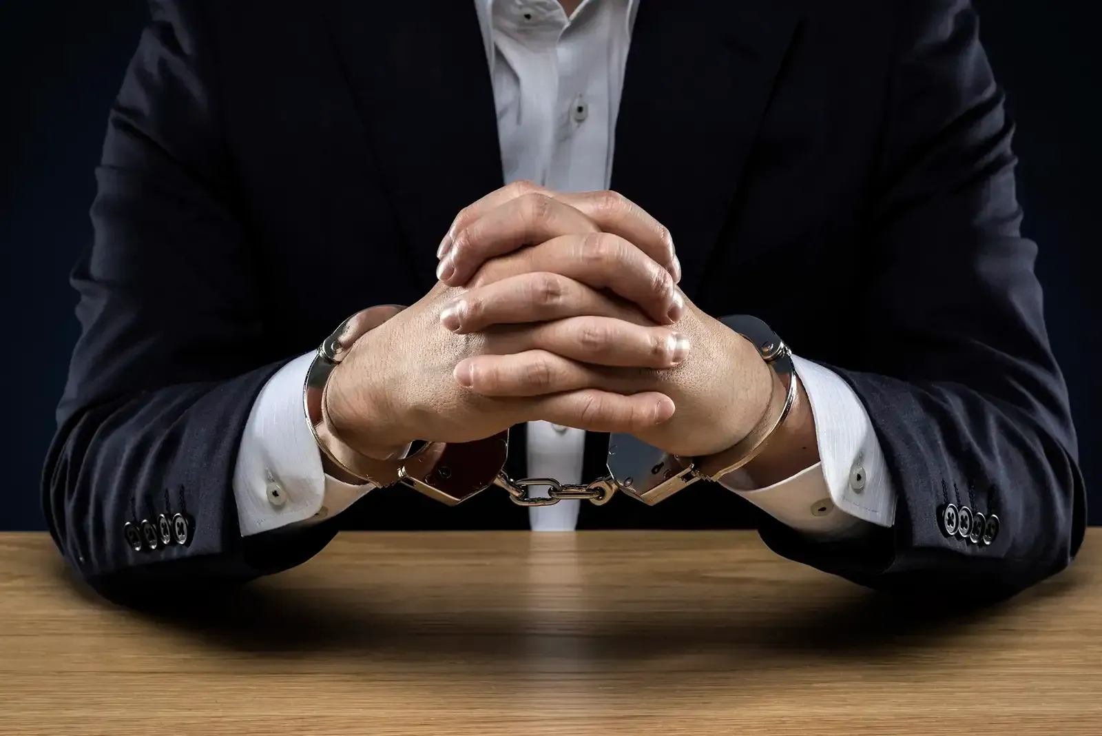 Man in suit with handcuffed hands on a wooden desk.