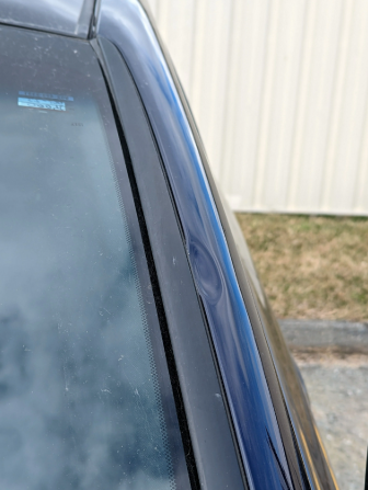 Close-up of a blue car’s windshield and side pillar with water spots, beside a beige wall.