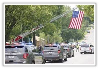 Police vehicles and fire truck ladder with American flag in street, trees in background.
