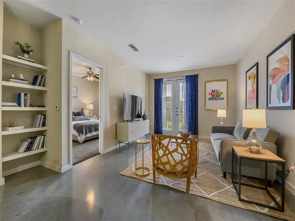 Living room and bedroom view in a modern apartment with a couch, chairs, shelving, and a bed.