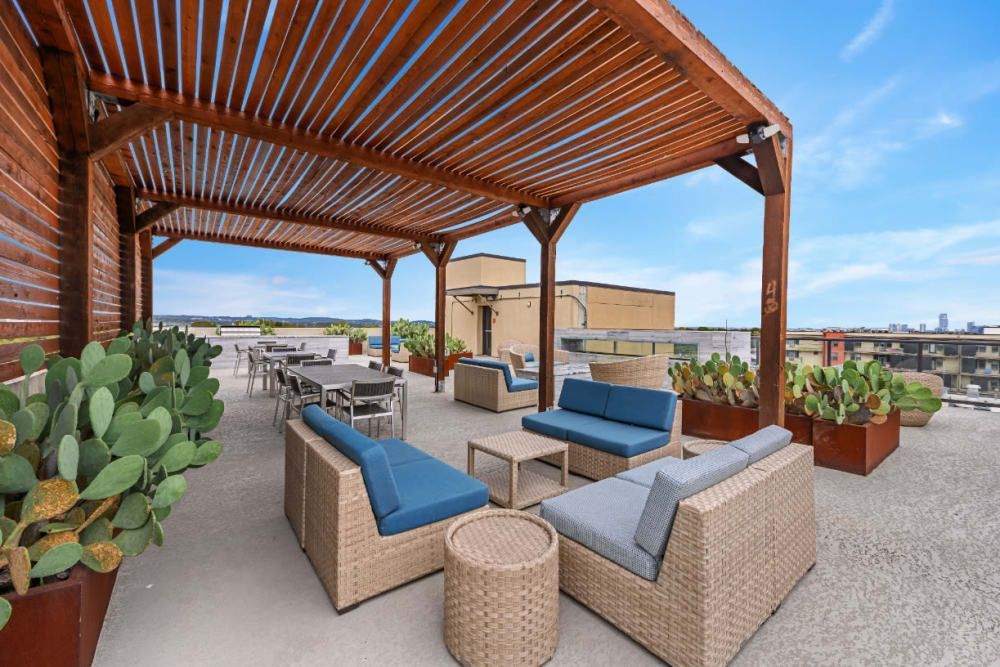 Rooftop patio with wicker seating, blue cushions, potted plants, and a wooden pergola overlooking the city.