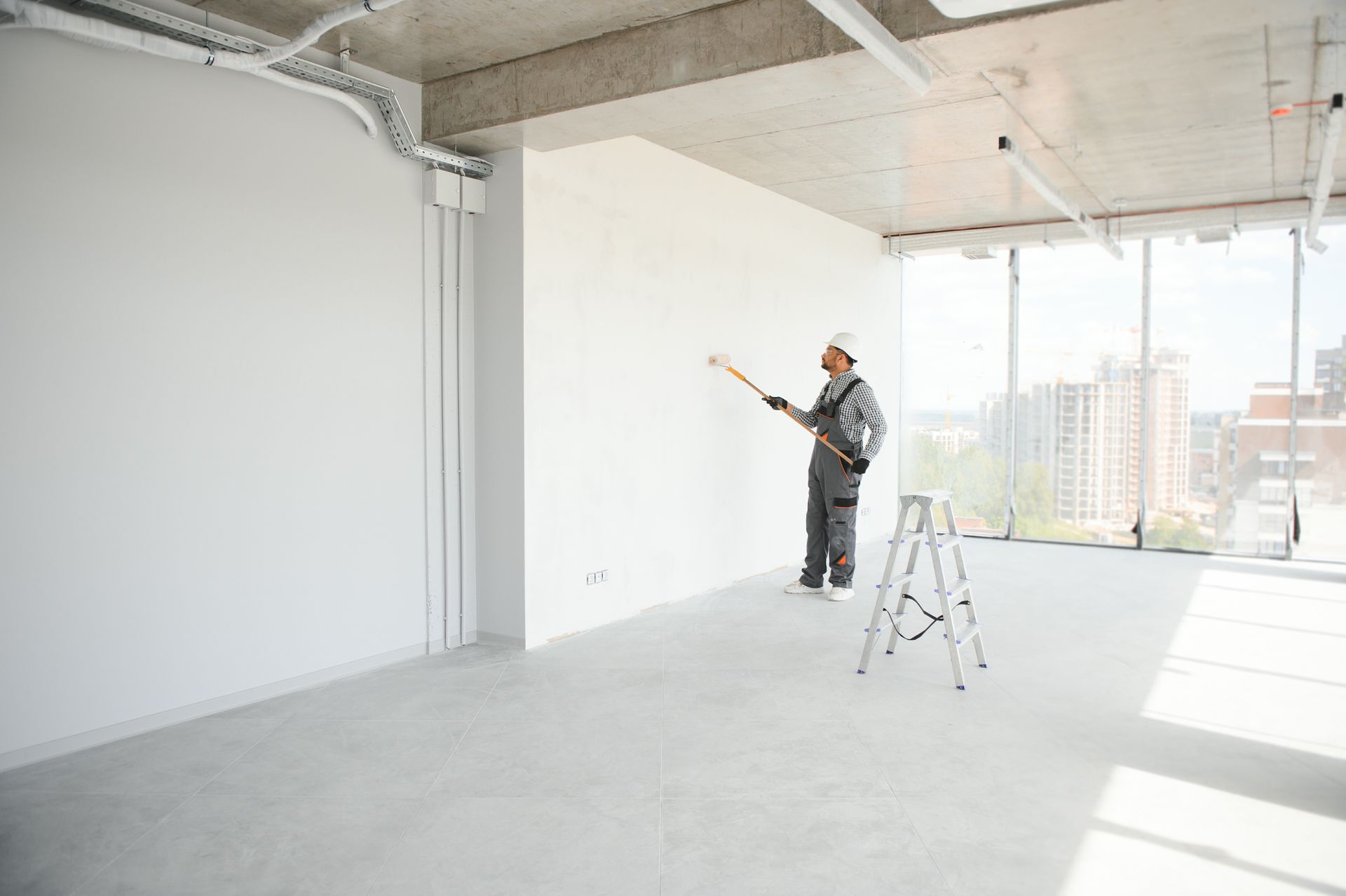 Worker painting a bright, empty room in a modern apartment with a ladder nearby