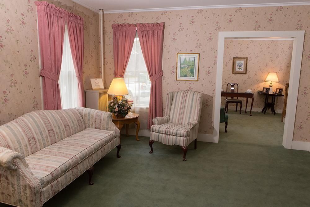 A furnished living room with floral wallpaper, pink curtains, sofa, armchair, and view into another room.