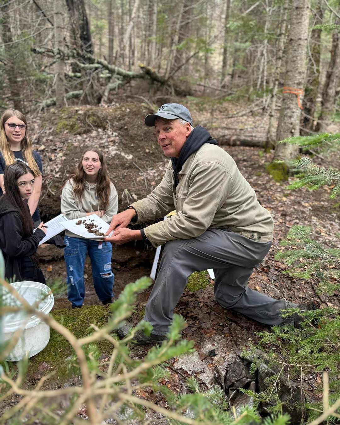 Man kneels, pointing at items in hand, surrounded by students in a wooded area, by a small water source.