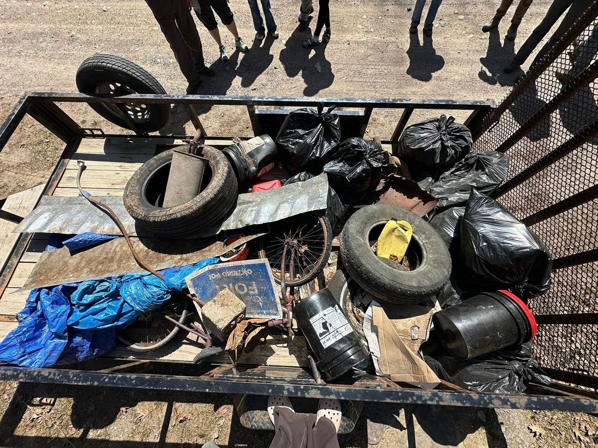 Cart filled with trash: tires, bags, metal, and debris. People's shadows overhead.