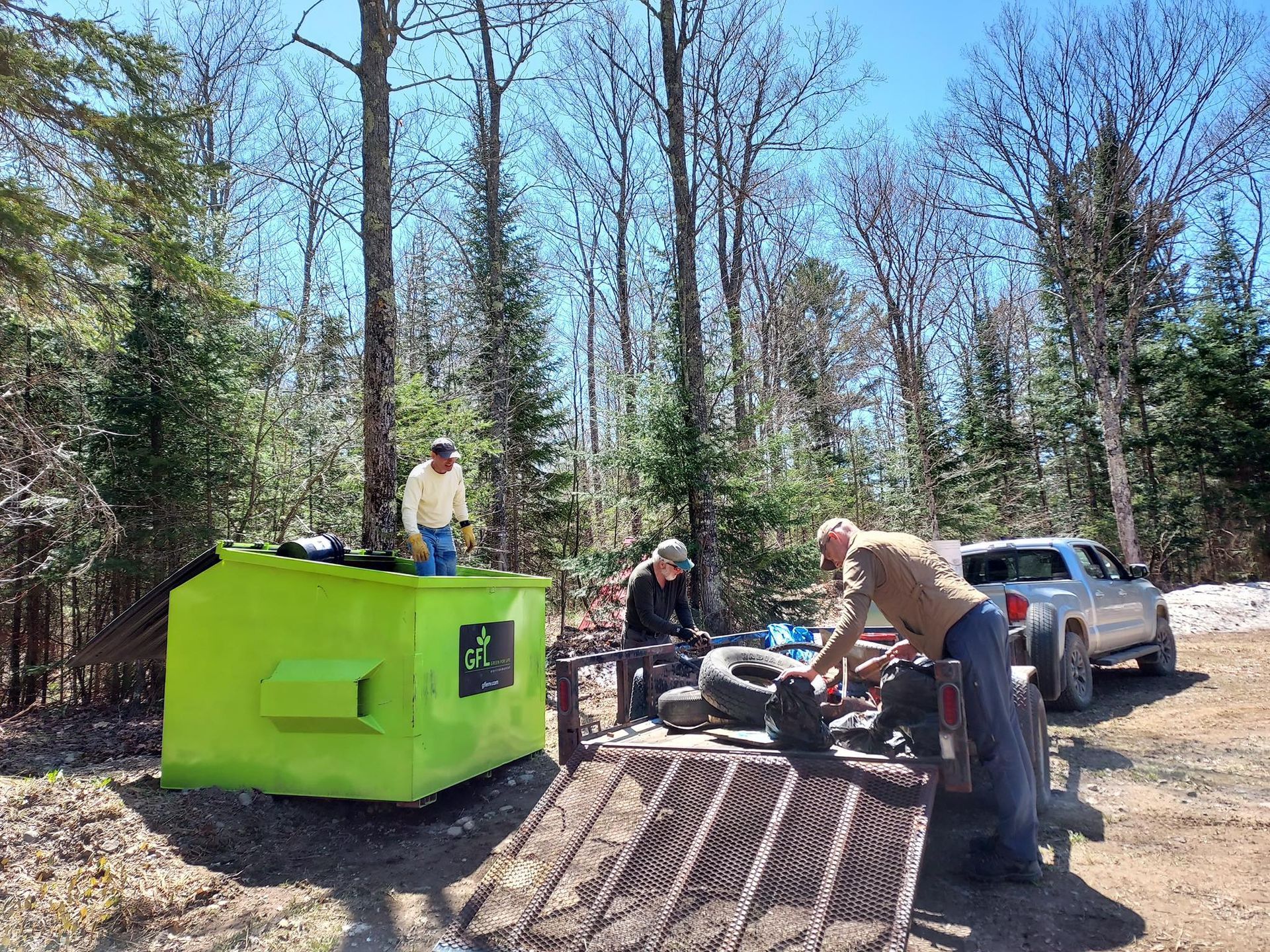 People working near a bright green structure and a trailer in a wooded area. A silver vehicle is parked nearby.