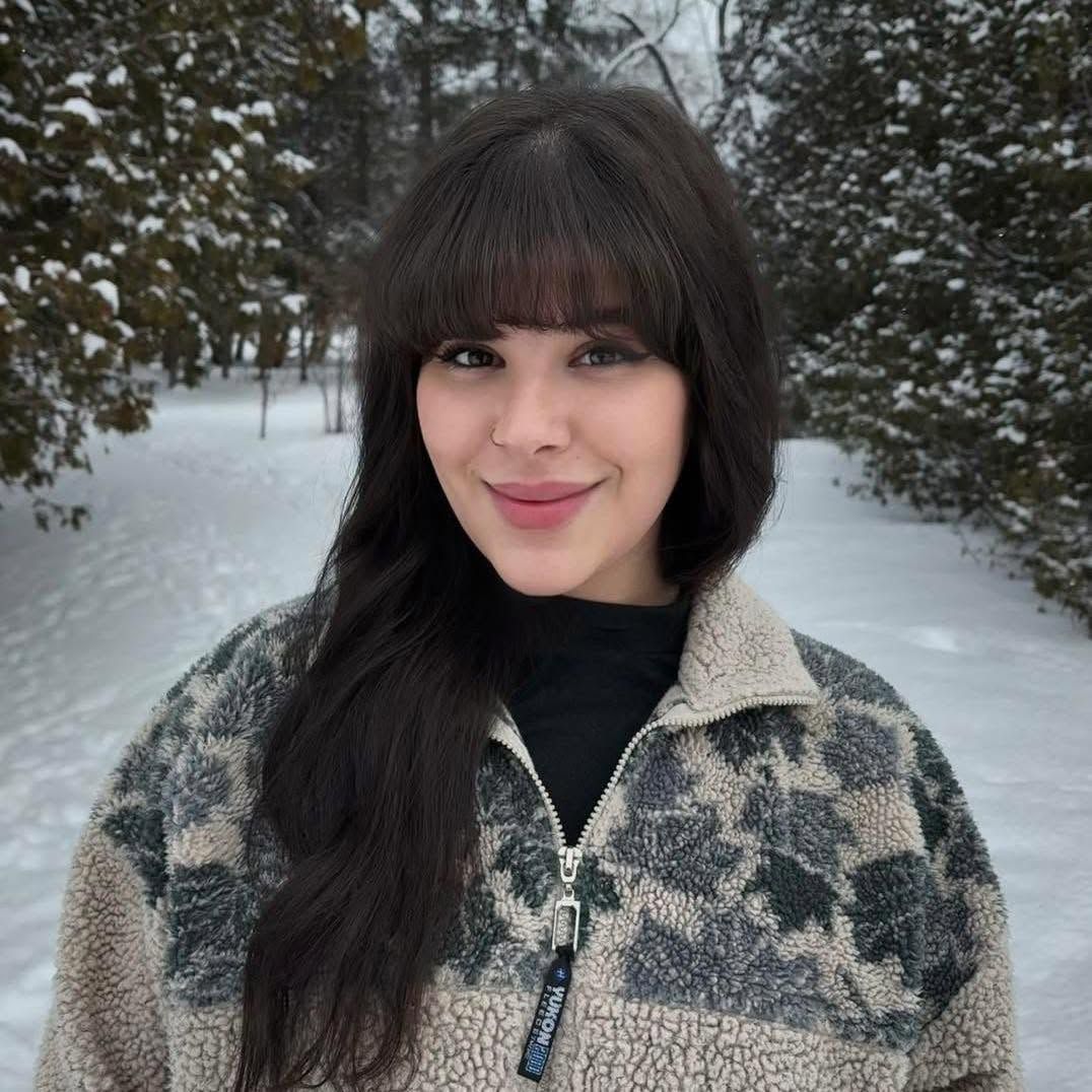 Woman smiling outdoors in a snowy forest, wearing a gray patterned jacket and black top