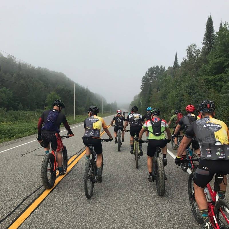 Group of cyclists riding on a paved road, mountains and fog in the background.
