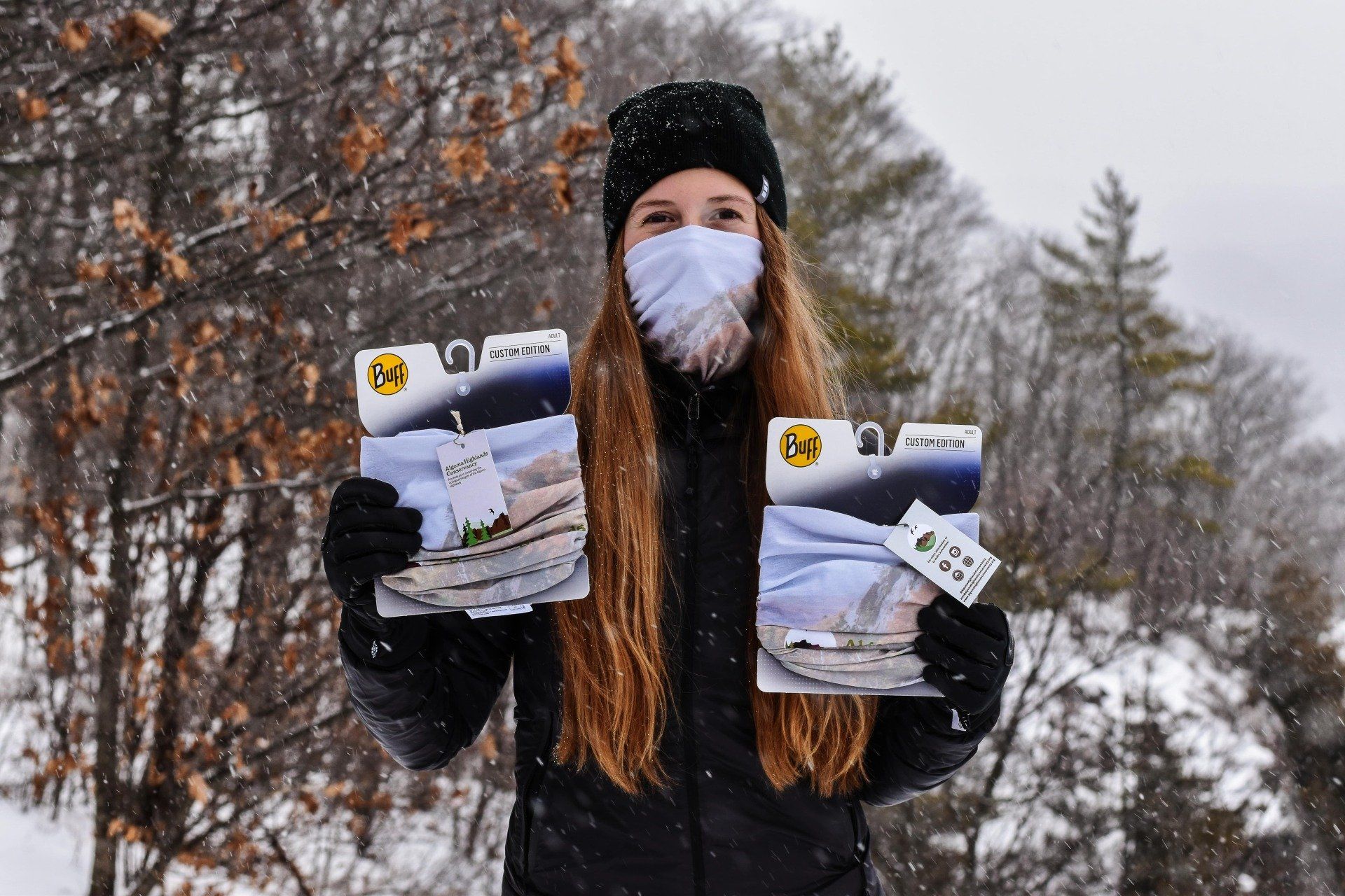 Person in winter gear holding two packaged items outdoors in a snowy forest, wearing a face mask.
