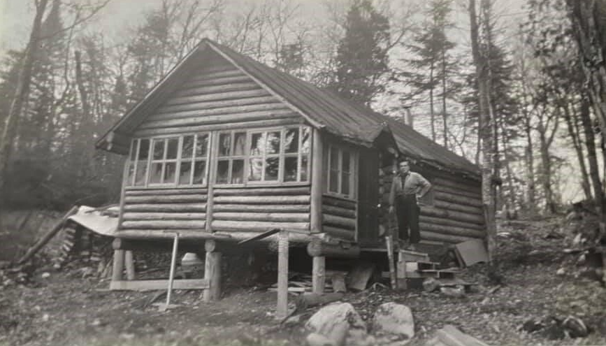 Log cabin in a wooded area with a person standing by the door.