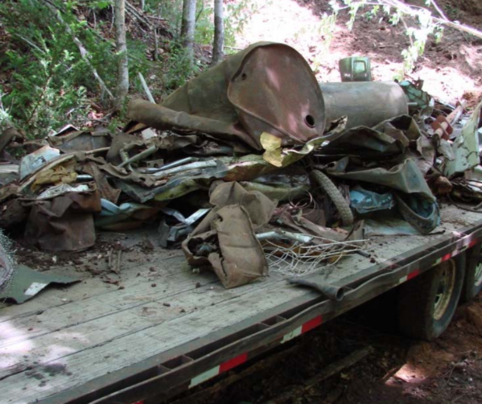 Rusty scrap metal piled on a flatbed trailer in a wooded outdoor area.