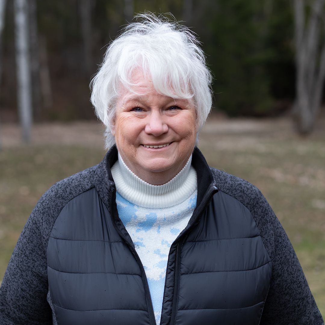 Smiling woman with short white hair wearing a dark jacket outdoors in a wooded area